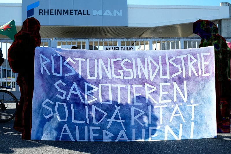 Mit den Menschen in Rojava solidarische Demonstrant_innen vor dem Rheinmetall Werk in Wien fordern auf einem Transparent: Rüstungsindustrie sabotieren, Solidarität aufbauen.